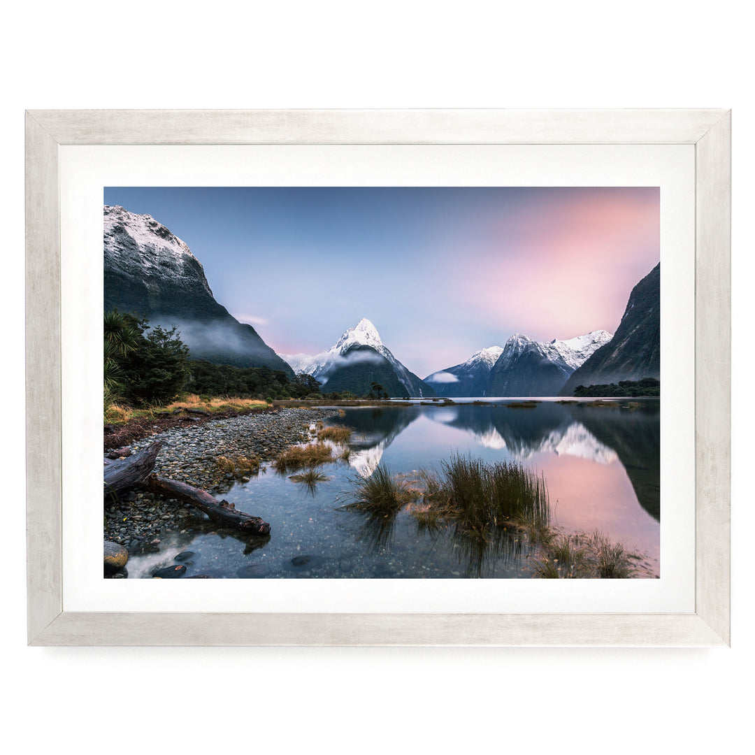 a serene landscape with a calm lake surrounded by snow-capped mountains, with a clear blue sky and a pink and orange sunset.