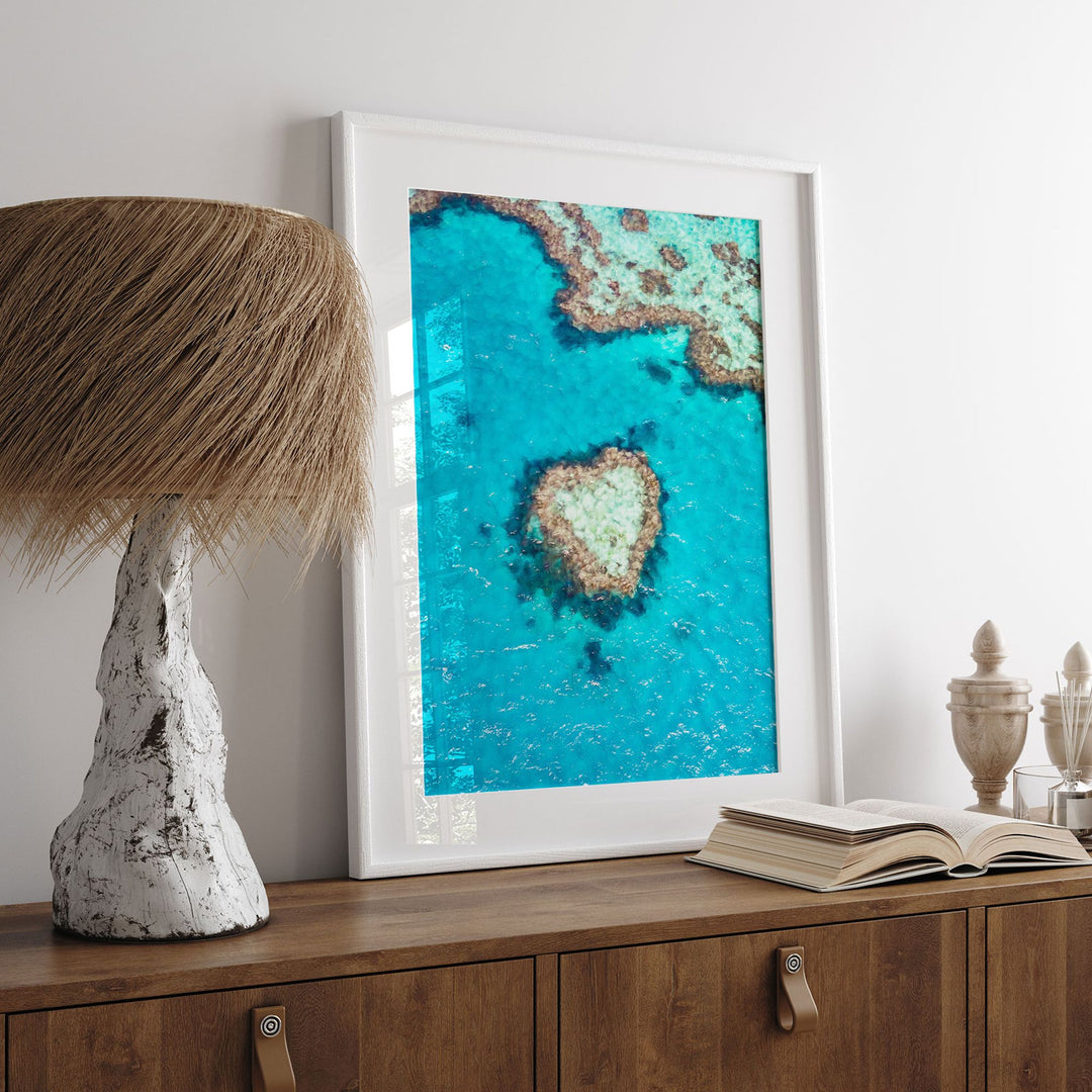 a framed photograph of a tropical island with a heart-shaped coral reef in the center, placed on a wooden dresser next to a lamp.