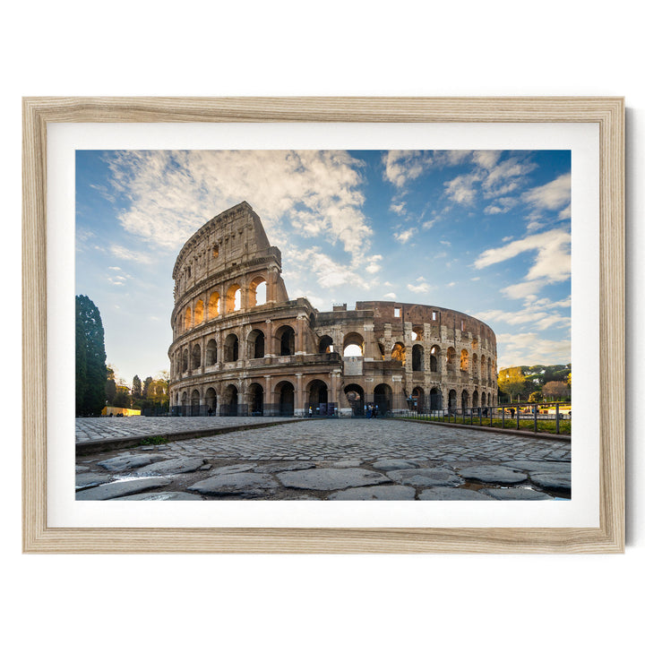 the Colosseum, an ancient amphitheater in Rome, Italy, with its distinctive oval shape and arches. The Colosseum is surrounded by a cobblestone courtyard, and the sky above is blue with some clouds. The photograph is framed and appears to be a piece of artwork or a printed image.