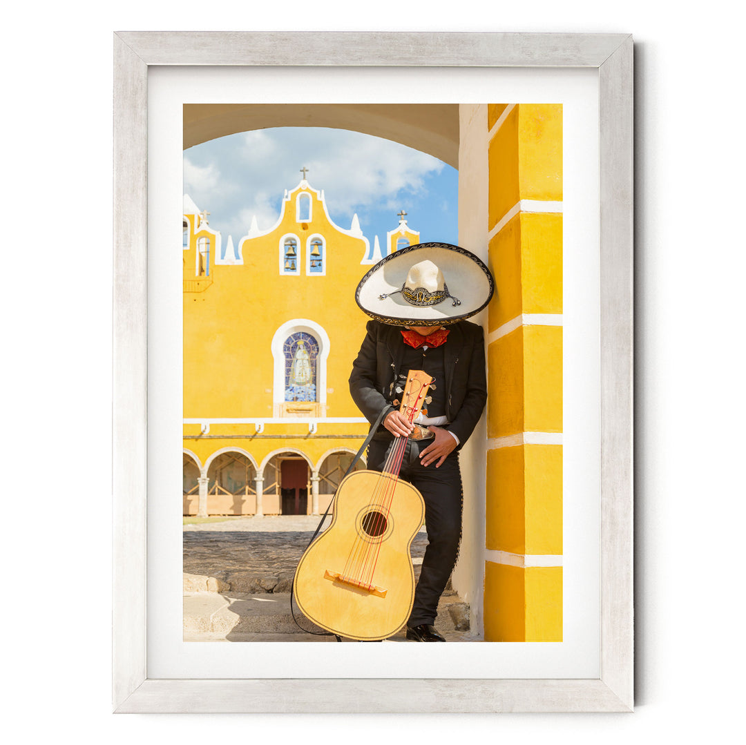 A person in a sombrero and traditional Mexican attire is playing a guitar in front of a yellow building with arched windows and a bell tower.