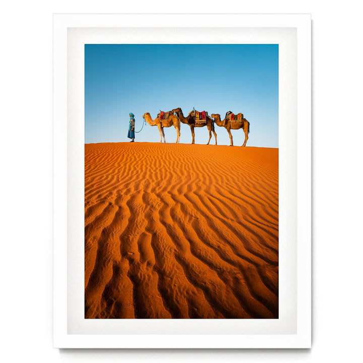 A person in blue attire leads three camels across a vast, undulating sand dune in a desert landscape.