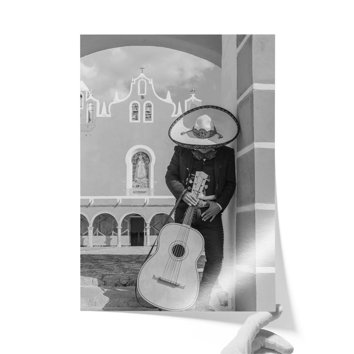 A person wearing a sombrero and holding a guitar is standing in front of a building with arched windows.