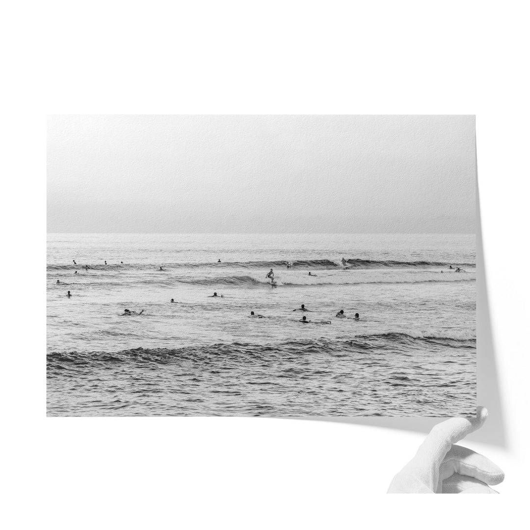 A black and white photograph of a group of people swimming and surfing in the ocean, with waves visible in the background.