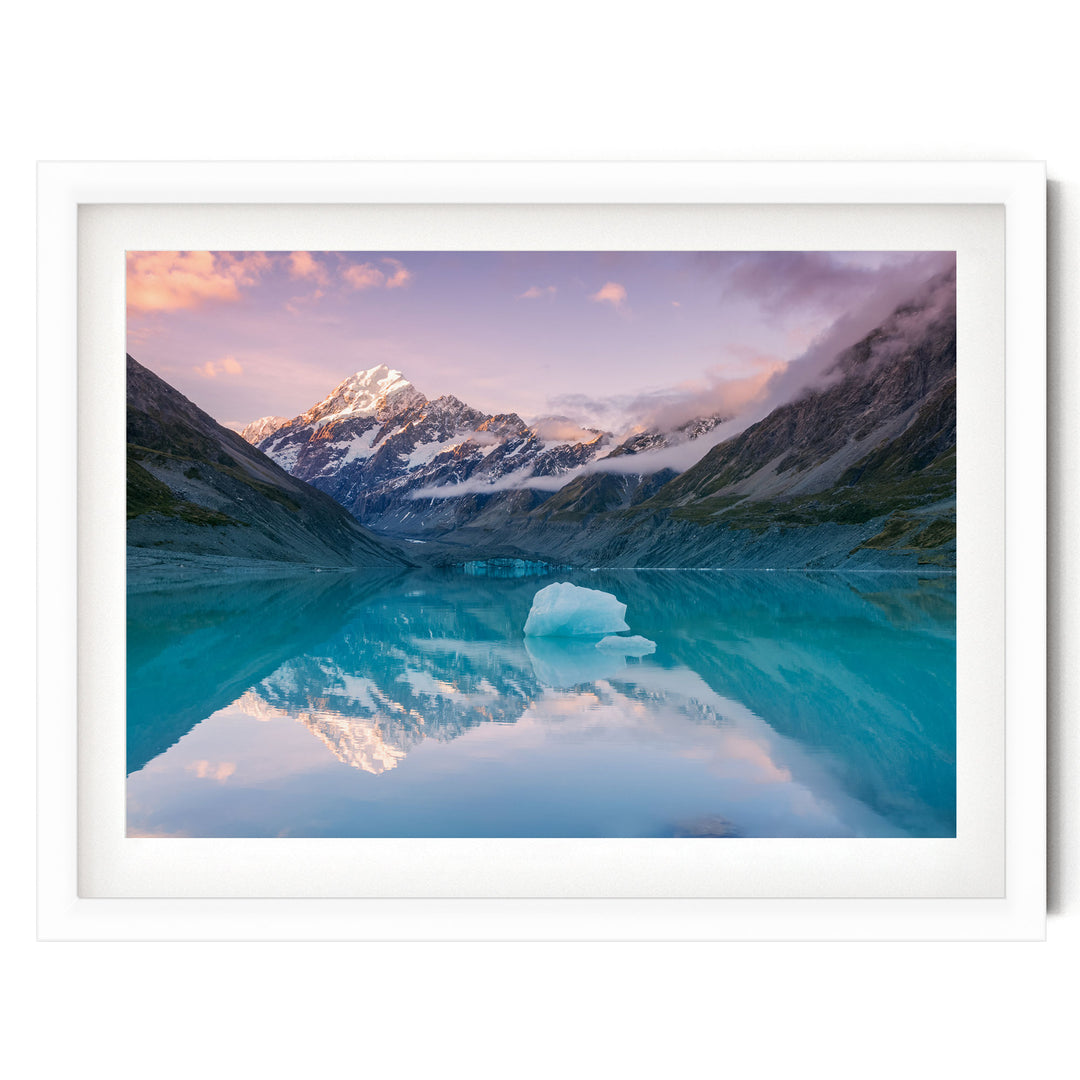 A breathtaking landscape featuring a serene lake surrounded by majestic snow-capped mountains, with a large iceberg floating on the calm waters. The sky above is a beautiful gradient of pink and purple hues, creating a stunning backdrop for the scene.