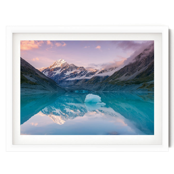 A breathtaking landscape featuring a serene lake surrounded by majestic snow-capped mountains, with a large iceberg floating on the calm waters. The sky above is a beautiful gradient of pink and purple hues, creating a stunning backdrop for the scene.