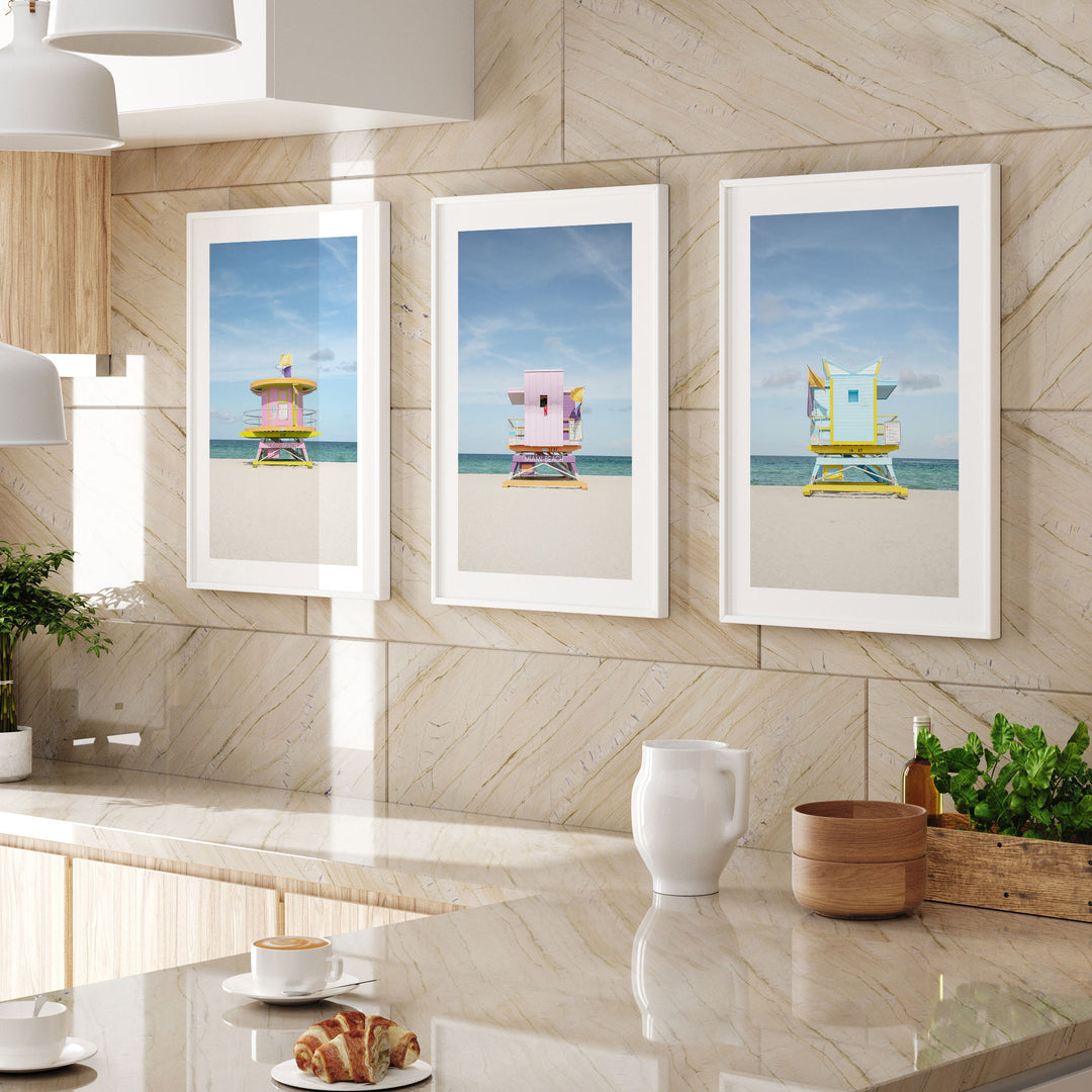 three framed beach lifeguard towers, each with a different color and design, displayed on a wall above a kitchen counter.