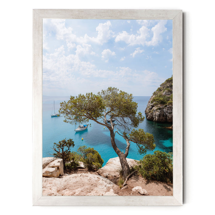 A scenic view of a rocky coastline with a large tree, a small boat, and a clear blue sky with white clouds.