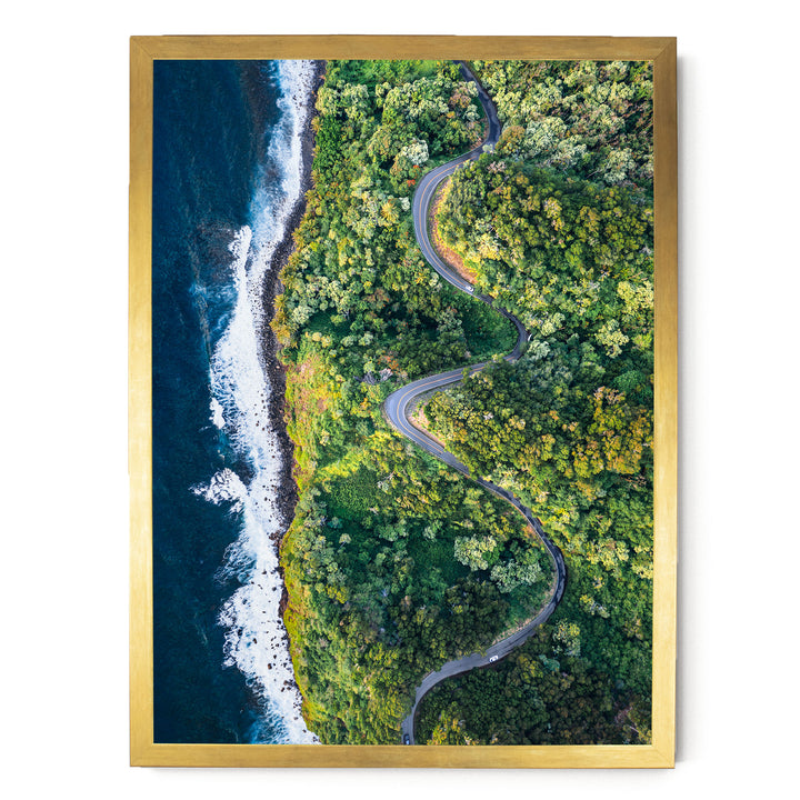 The image is an aerial view of a winding road cutting through a lush, green forest. The road curves gently around the edge of a cliff, with the ocean visible on the left side. The surrounding landscape is dense with trees and vegetation, creating a serene and picturesque scene.