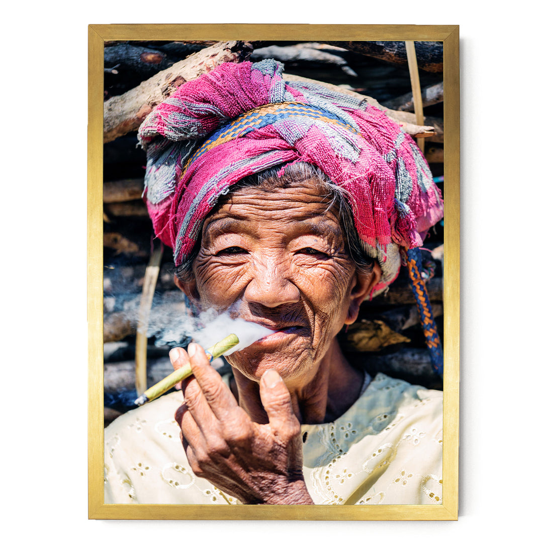 An elderly woman wearing a colorful headscarf and holding a lit cigarette, with a pile of wood in the background.