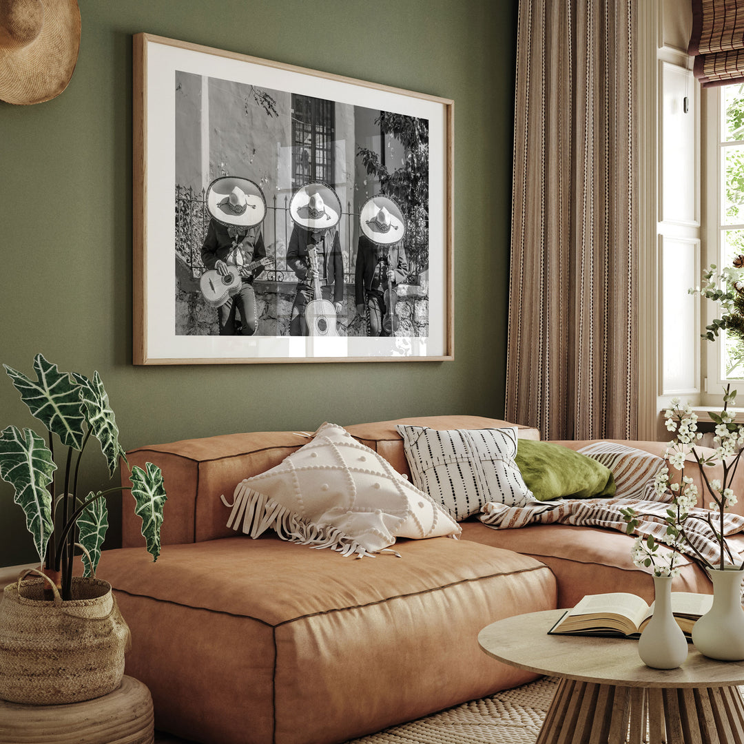 a cozy living room with a brown leather sofa adorned with throw pillows and a green throw blanket. Above the sofa, a framed black and white photograph of three people playing musical instruments is displayed on the wall. The room is further decorated with potted plants, vases, and a wooden coffee table, creating a warm and inviting atmosphere.