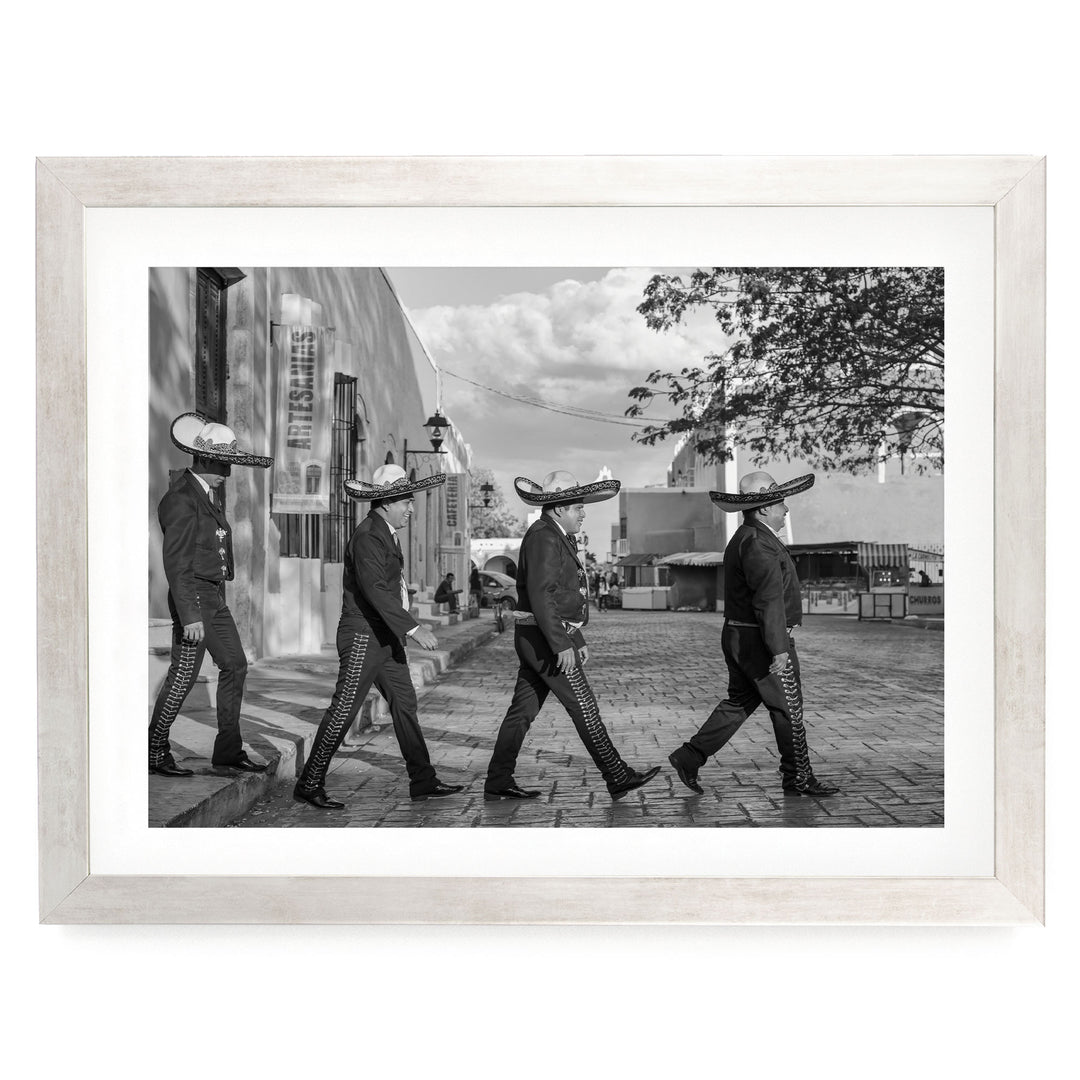 A group of men wearing sombreros are walking down a street, with a building and trees in the background.