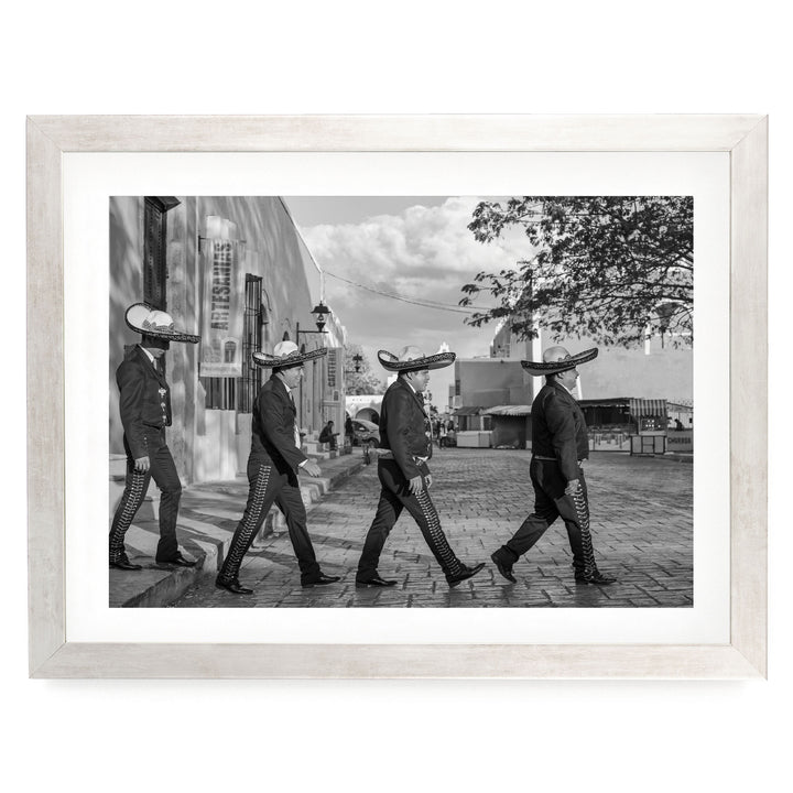 A group of men wearing sombreros are walking down a street, with a building and trees in the background.