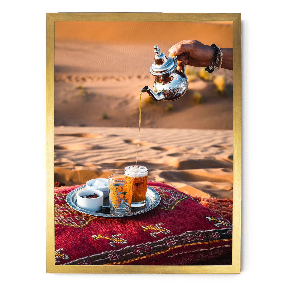 A hand is pouring tea from a silver teapot into two glasses filled with tea, set on a red and gold patterned cushion or pillow.