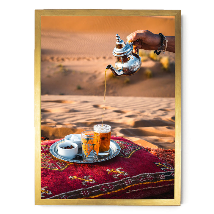A hand is pouring tea from a silver teapot into two glasses filled with tea, set on a red and gold patterned cushion or pillow.