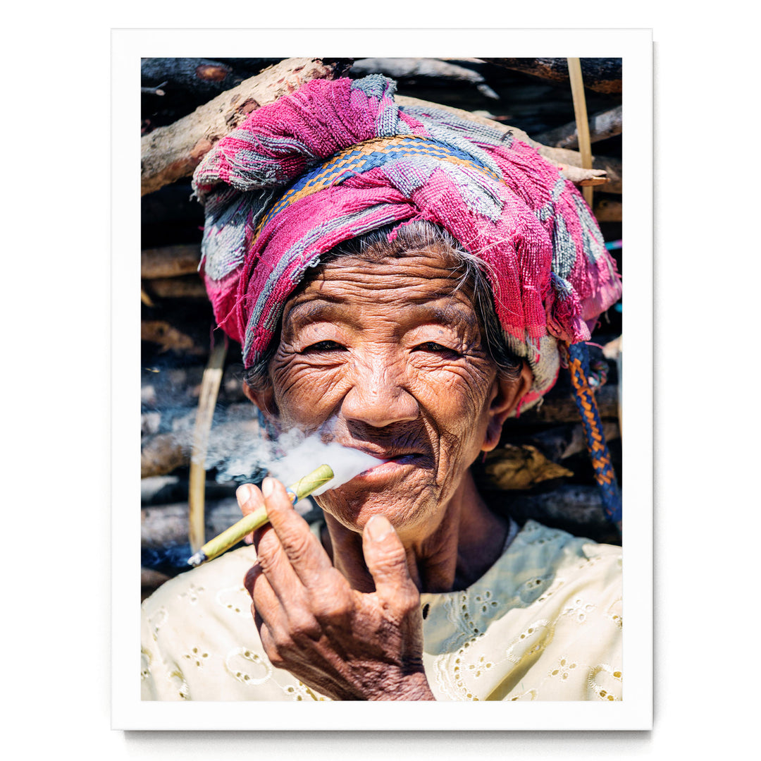 An elderly woman wearing a colorful headscarf and holding a lit cigarette, with a pile of wood in the background.