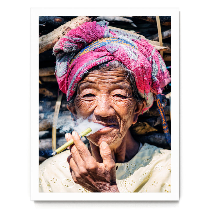 An elderly woman wearing a colorful headscarf and holding a lit cigarette, with a pile of wood in the background.