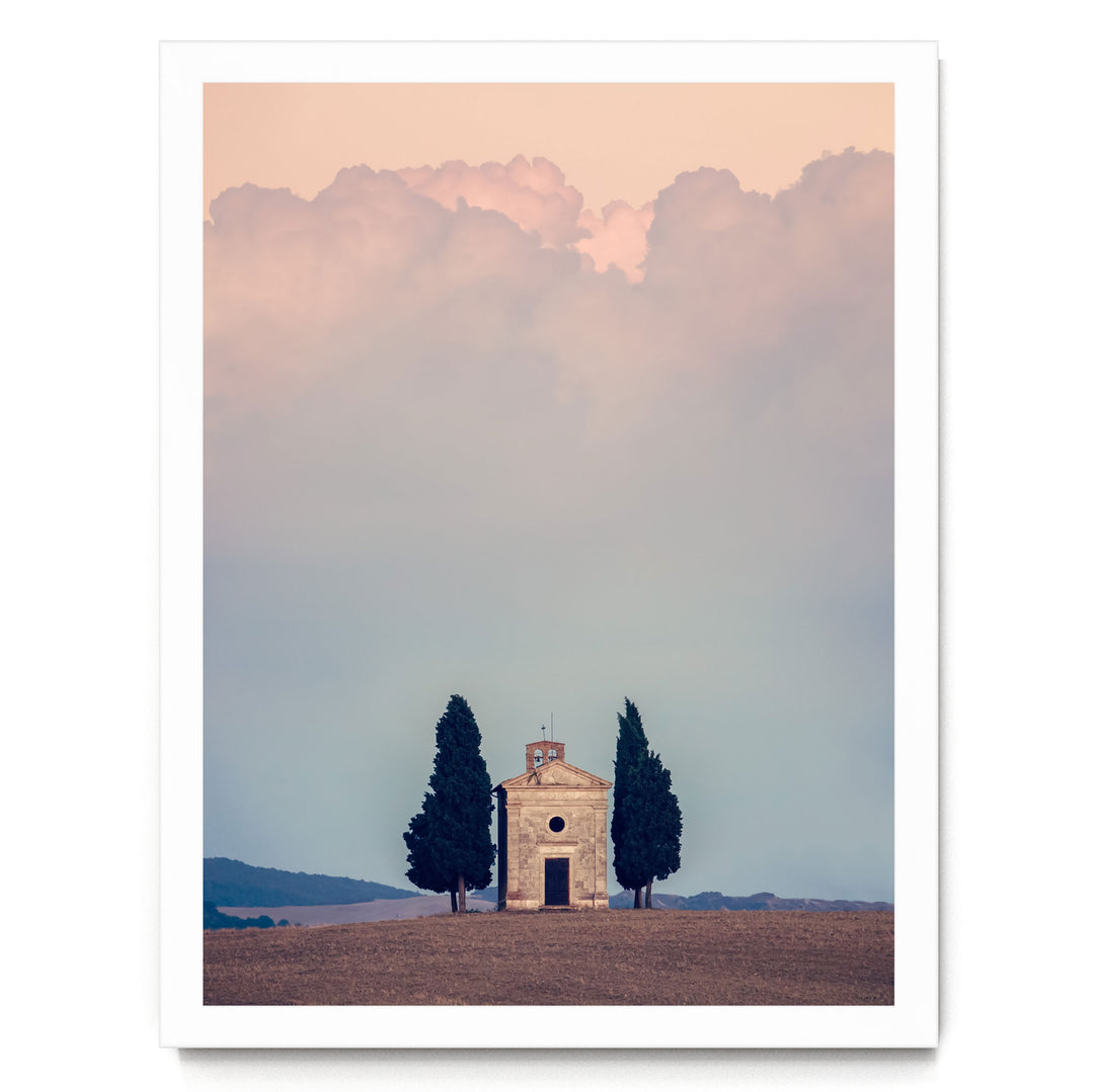 A small stone church with a bell tower stands in a field, surrounded by two tall trees, with a cloudy sky in the background.