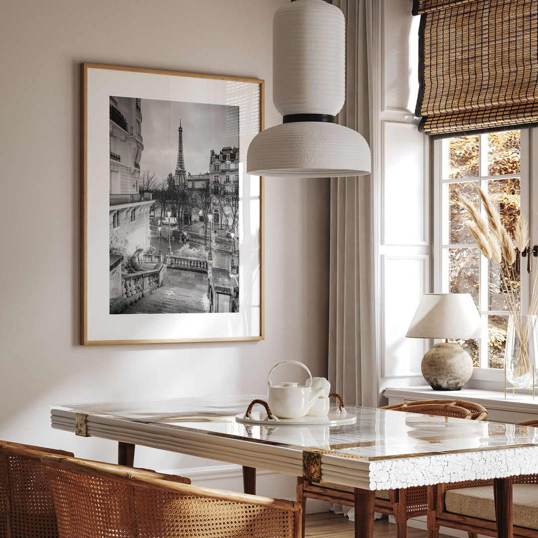 a dining room with a large framed picture of the Eiffel Tower hanging on the wall above a dining table with a white tablecloth. The table is set with a teapot and a vase, and there are chairs around the table. The room has a modern and elegant design, with a large window providing natural light and a view of the outdoors.