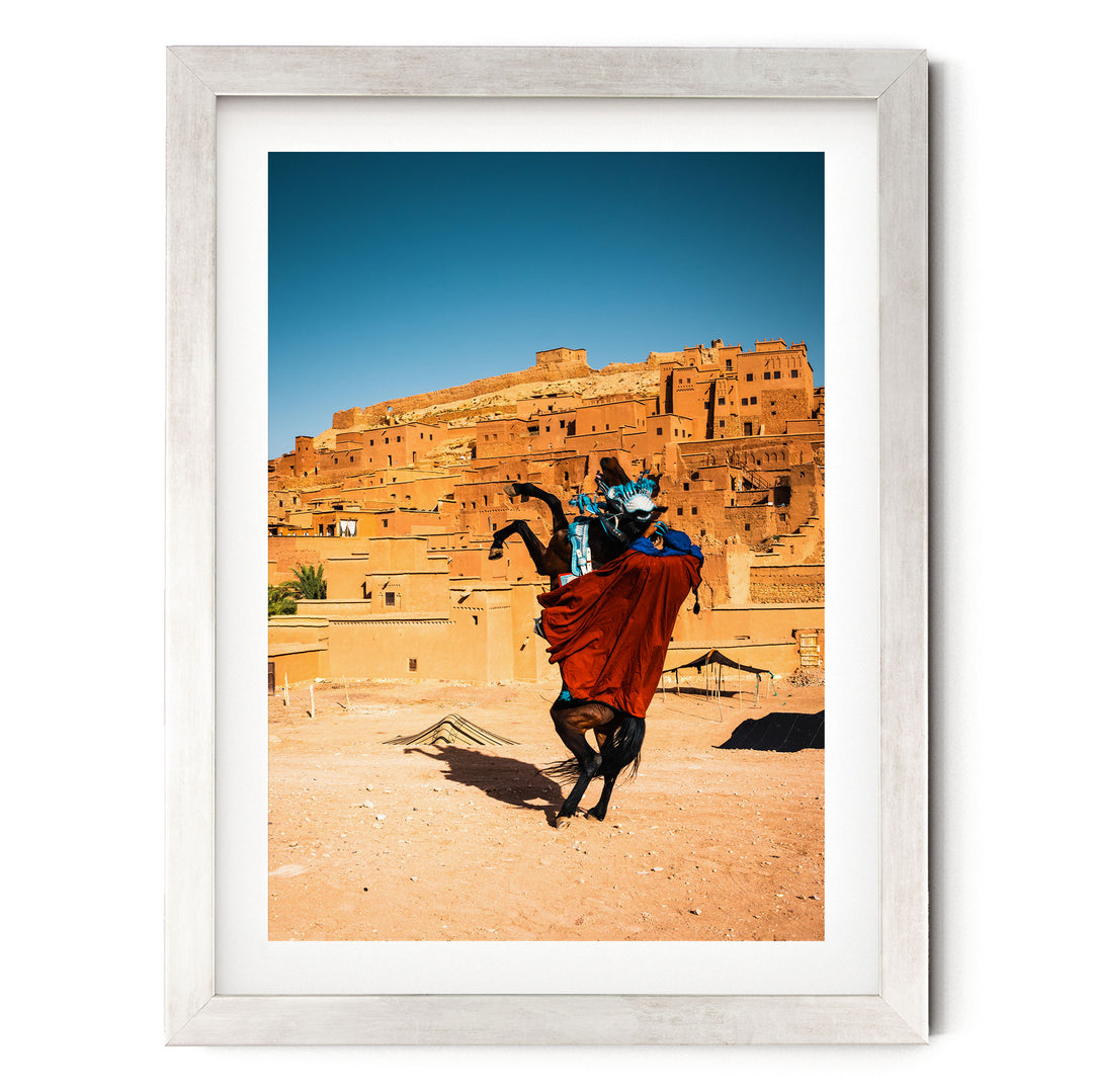 A person riding a horse in a desert-like setting, with a large, multi-story structure in the background.