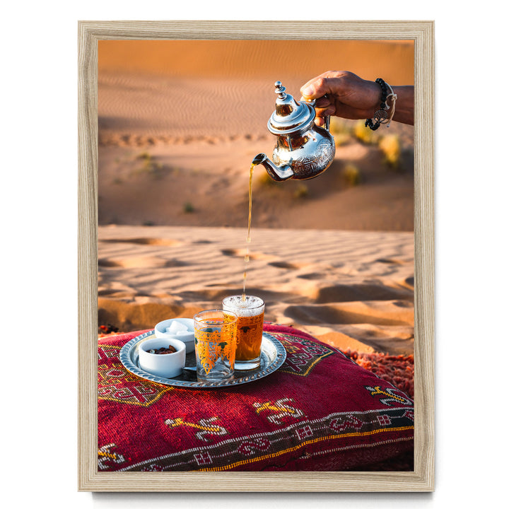 A hand pours tea from a silver teapot into two glasses on a tray, set on a red and gold patterned cushion, with a desert landscape in the background.