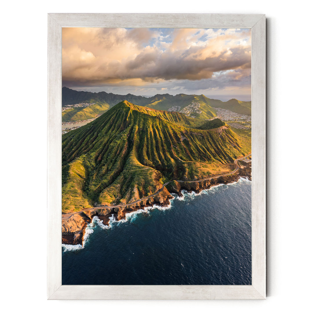 a stunning mountainous landscape with a winding road cutting through the rugged terrain. The mountains are covered in lush green vegetation, and the ocean is visible in the background. The sky is filled with clouds, creating a dramatic backdrop for the scene. The image is framed, suggesting it is a piece of art or a photograph.