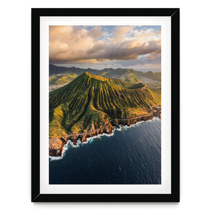 a mountainous coastline with a steep, jagged peak overlooking the ocean. The sky is filled with clouds, creating a dramatic backdrop for the scene. The photograph is framed and appears to be a high-quality image of a natural landscape.
