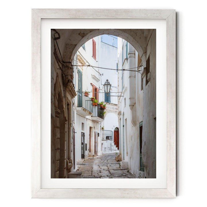A narrow cobblestone street lined with white buildings and colorful shutters, leading to a small balcony with potted plants.