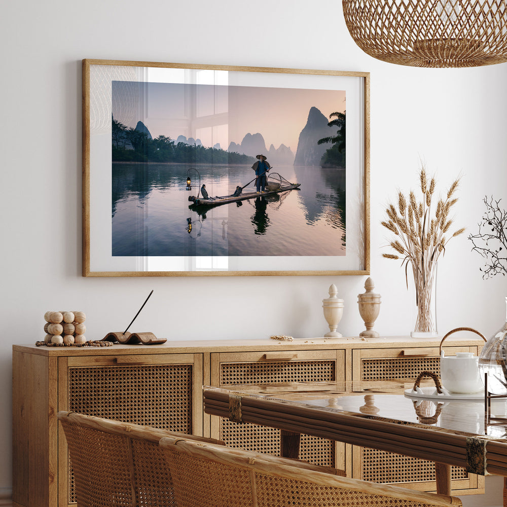 a serene scene of a person on a boat on a calm lake, with mountains in the background. The photograph is framed and hung on a wall above a wooden cabinet, which has a vase of flowers and a bowl on top. The overall setting appears to be a peaceful and tranquil indoor space.