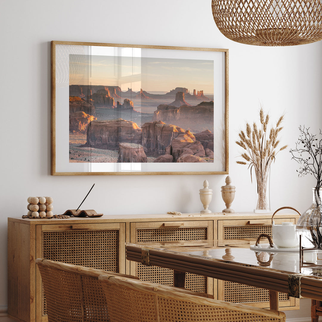 a large framed picture of a desert landscape hanging on a wall above a wooden cabinet with drawers. The cabinet has a glass top and is situated in a dining area with a table and chairs. The overall setting appears to be a cozy and inviting space, with a warm and natural aesthetic.