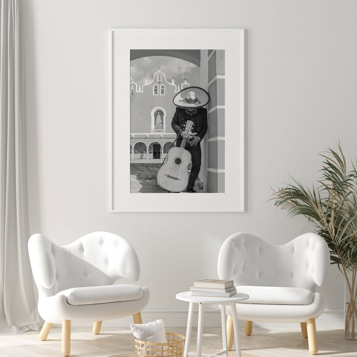 a framed black and white photograph of a person playing a guitar, standing in front of a building with arches. The photograph is hung on a white wall above two white armchairs and a small white table.