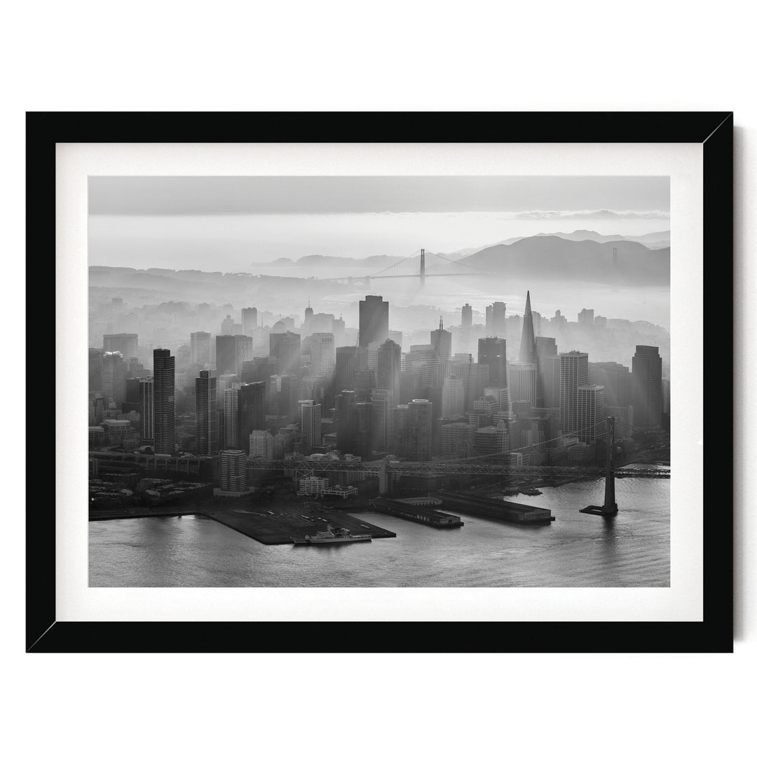 A black and white aerial view of a densely populated city skyline, with tall buildings and skyscrapers stretching out across the frame. The city is situated near a body of water, with a bridge visible in the distance. The sky above the city is hazy, creating a dramatic and atmospheric effect.