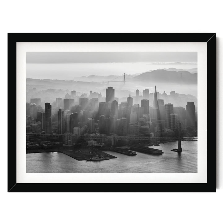 A black and white aerial view of a densely populated city skyline, with tall buildings and skyscrapers stretching out across the frame. The city is situated near a body of water, with a bridge visible in the distance. The sky above the city is hazy, creating a dramatic and atmospheric effect.