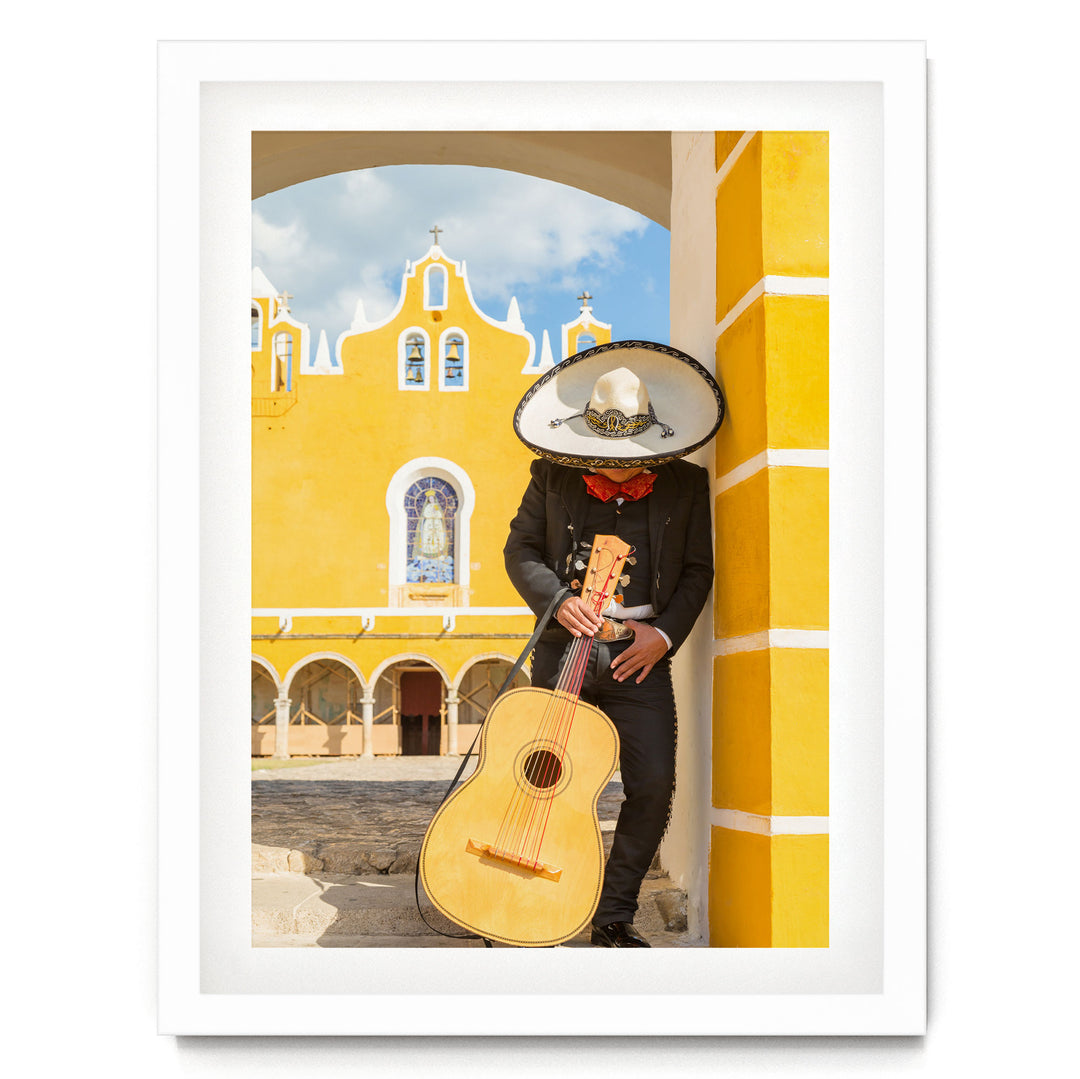 A person in a sombrero and traditional Mexican attire is playing a guitar in front of a yellow building with arched windows and a cross on top.