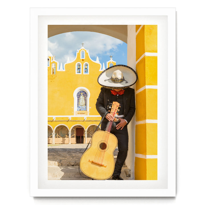 A person in a sombrero and traditional Mexican attire is playing a guitar in front of a yellow building with arched windows and a cross on top.