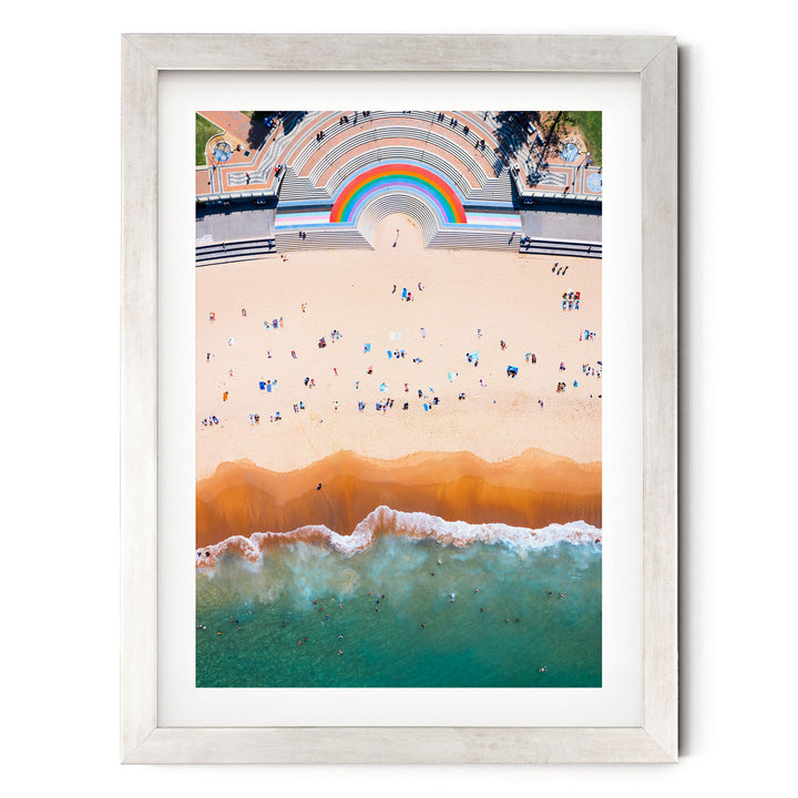 The image is an aerial view of a beach with a crowd of people enjoying the sunny day. The beach has a vibrant, colorful atmosphere with a rainbow arching over the top. The ocean is a beautiful shade of turquoise, and the sky is a clear blue. The image is framed and appears to be a collage of multiple images, creating a visually striking and dynamic scene.