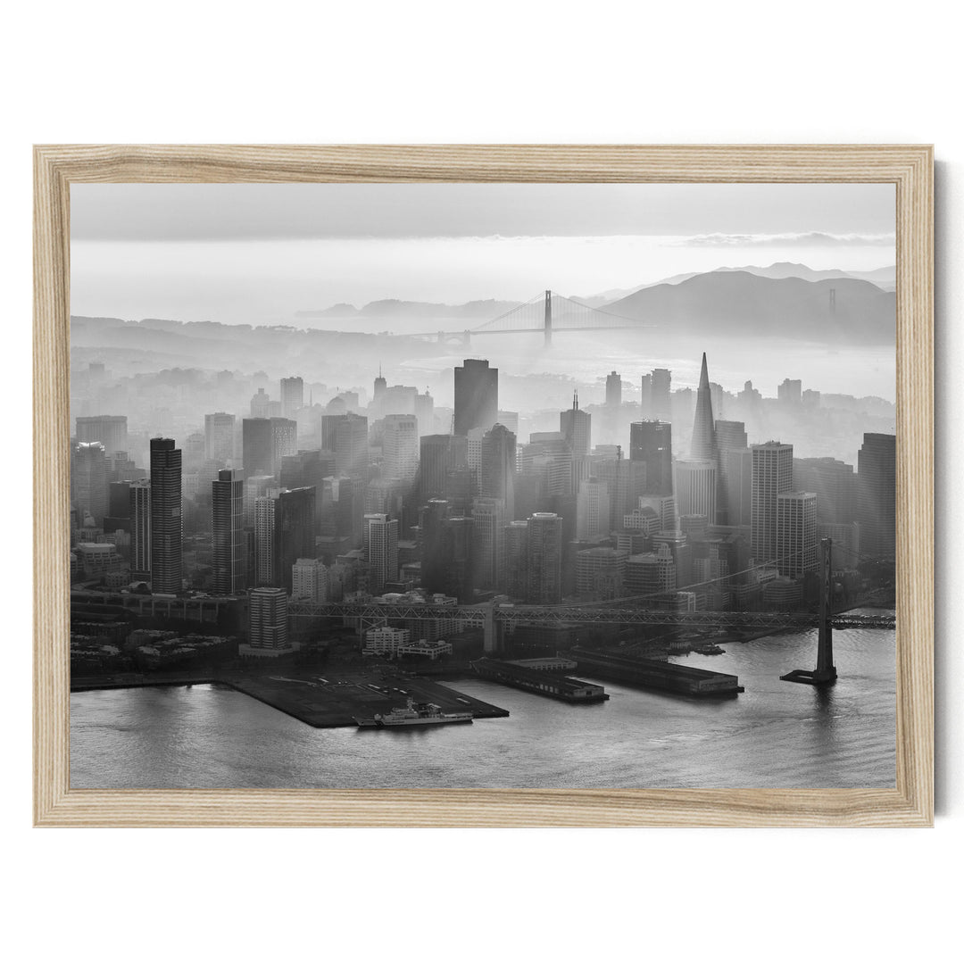 A black and white aerial view of a densely populated city skyline, with the Golden Gate Bridge and foggy mountains in the background.
