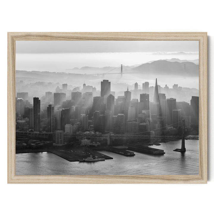 A black and white aerial view of a densely populated city skyline, with the Golden Gate Bridge and foggy mountains in the background.
