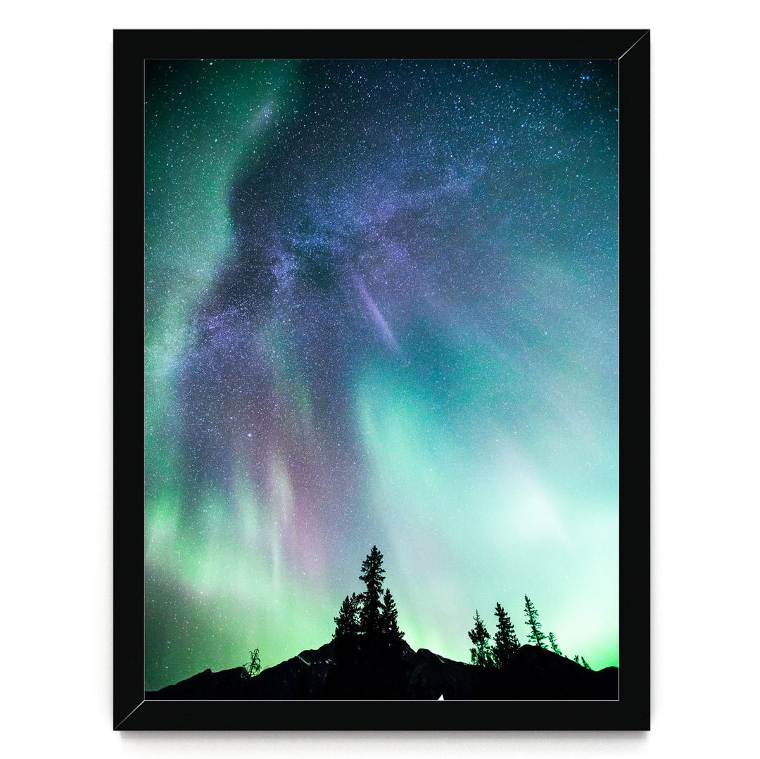 a stunning night sky with a vibrant display of the Aurora Borealis, or Northern Lights, illuminating the surrounding landscape. The silhouettes of trees can be seen in the foreground, adding a sense of depth and scale to the scene. The colors in the sky are a mix of green, blue, purple, and pink, creating a mesmerizing and otherworldly atmosphere.