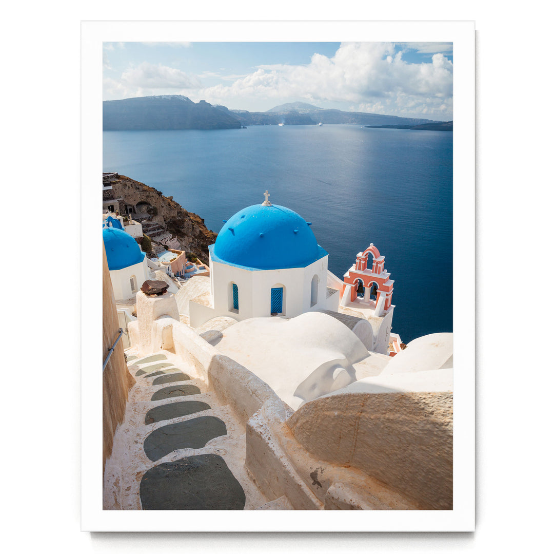 a picturesque view of a Greek island, with white buildings featuring blue domes and red roofs overlooking the deep blue sea. The sky is clear and blue, creating a serene and idyllic atmosphere.