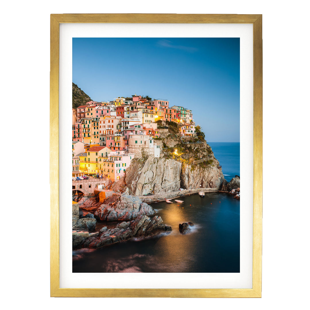 a picturesque coastal town nestled on a cliff, with its buildings illuminated by warm lighting against a backdrop of a clear blue sky and the calm sea.
