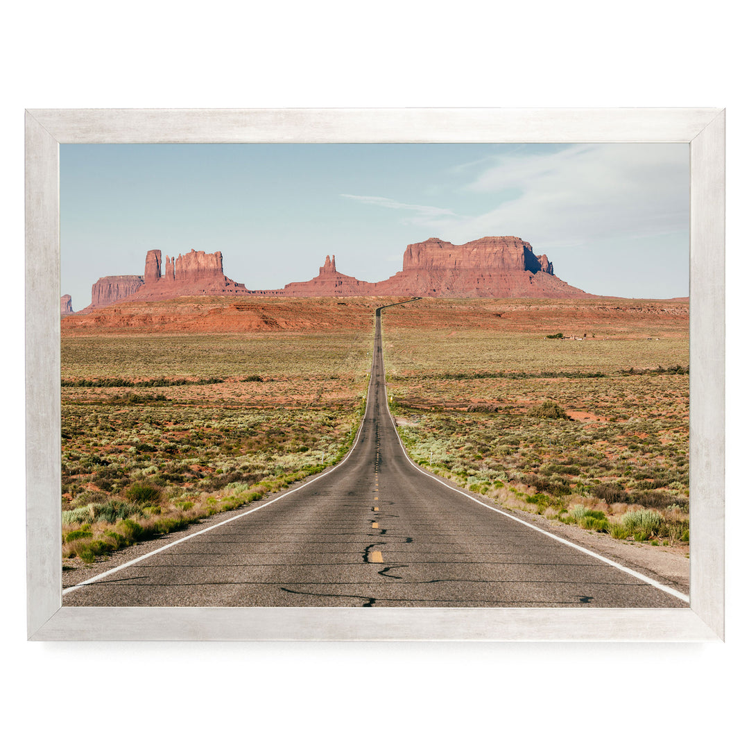 A long, straight road stretches out into the distance, leading towards a series of red rock formations in the background.