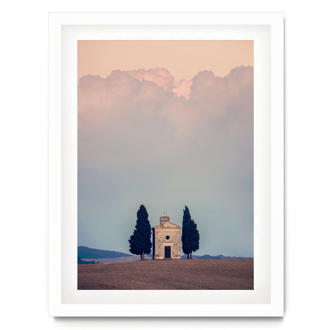 A white framed picture depicting a small stone church with two tall cypress trees in the foreground, set against a cloudy sky.