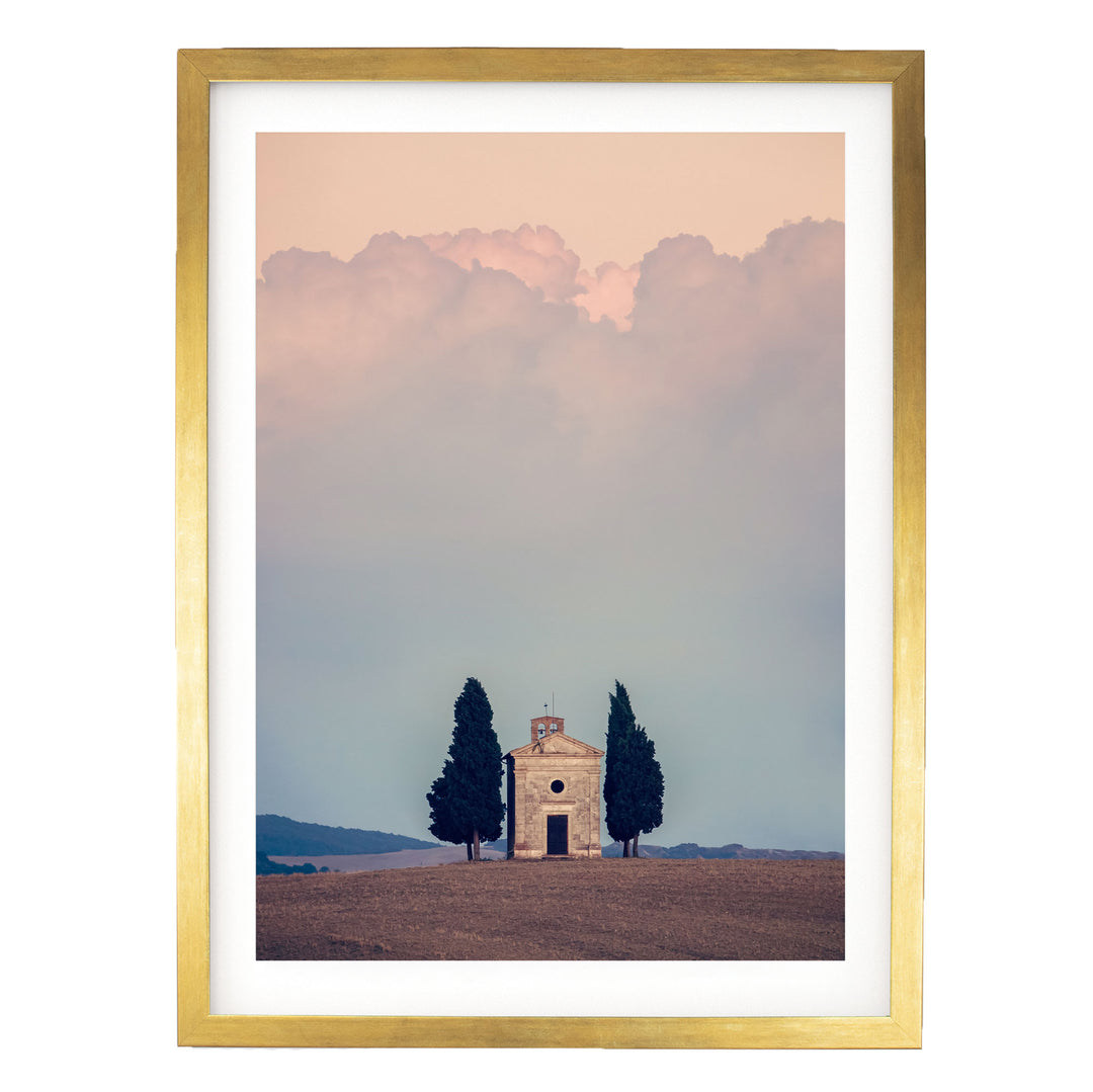 A small white church with a tower sits in a field, surrounded by two tall trees, with a cloudy sky in the background.
