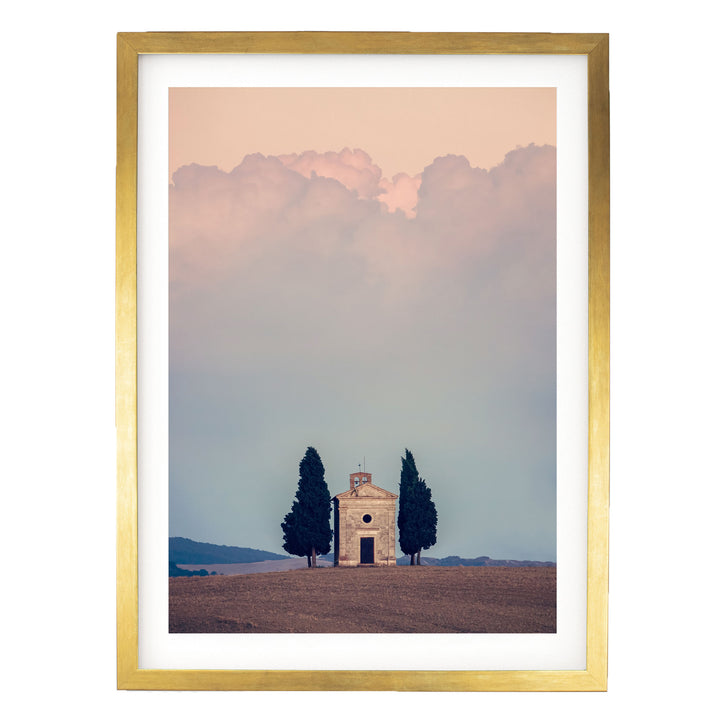 A small white church with a tower sits in a field, surrounded by two tall trees, with a cloudy sky in the background.