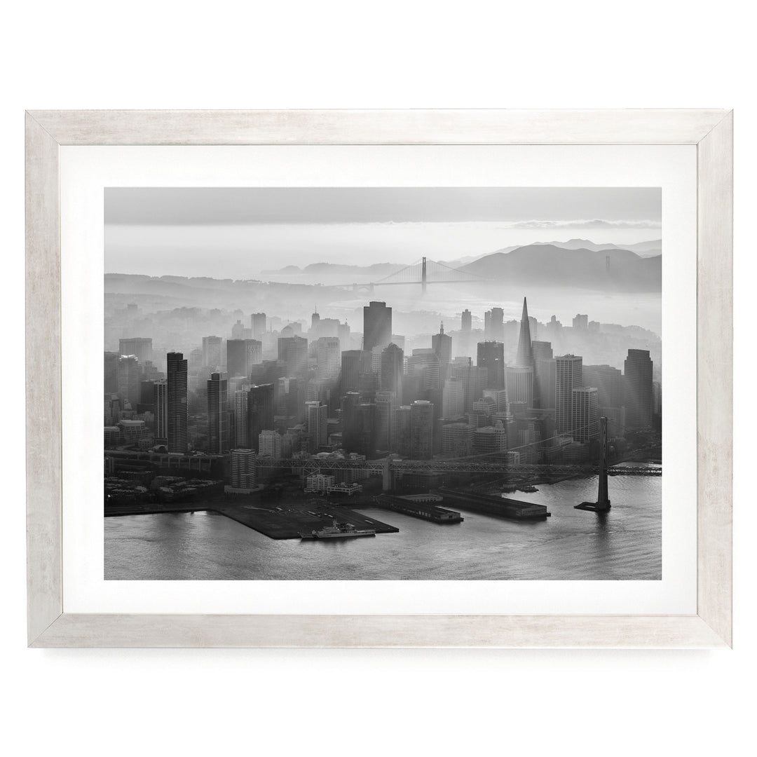 A black and white aerial view of a densely populated city skyline, with tall buildings and skyscrapers stretching out across the frame. The city is situated near a body of water, with a bridge visible in the distance. The sky above the city is hazy, creating a dramatic and atmospheric effect.