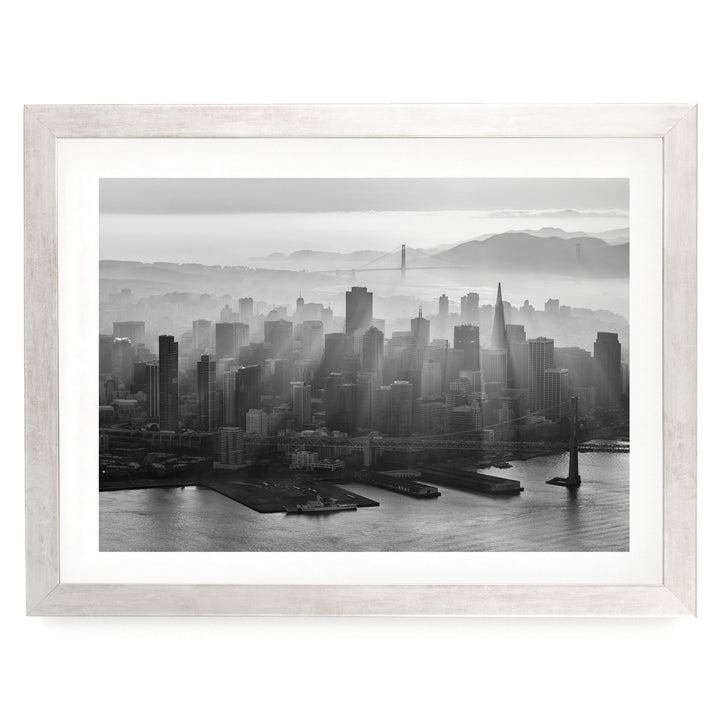A black and white aerial view of a densely populated city skyline, with tall buildings and skyscrapers stretching out across the frame. The city is situated near a body of water, with a bridge visible in the distance. The sky above the city is hazy, creating a dramatic and atmospheric effect.