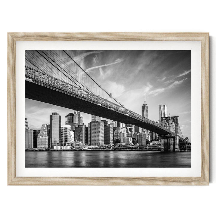 A black and white photograph of the Brooklyn Bridge spanning the East River, with the city skyline of New York City visible in the background.