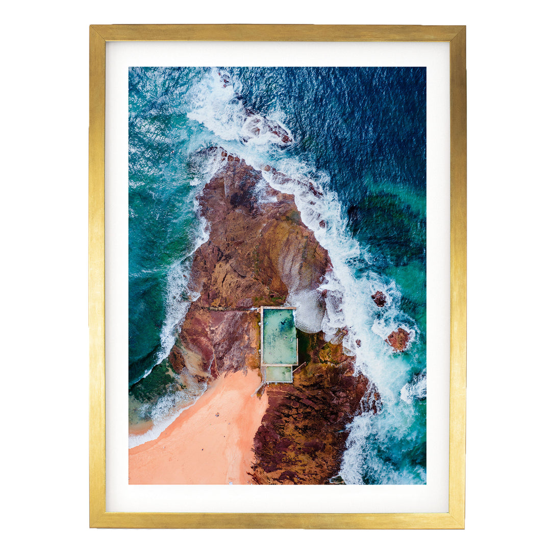 The image is an aerial view of a beach with a swimming pool, surrounded by rocky cliffs and crashing waves.