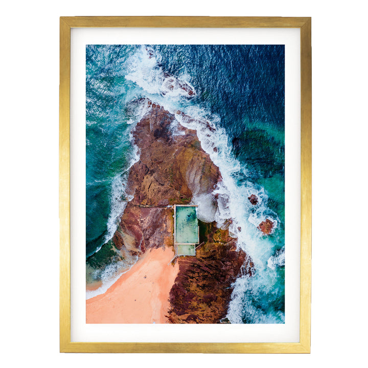 The image is an aerial view of a beach with a swimming pool, surrounded by rocky cliffs and crashing waves.