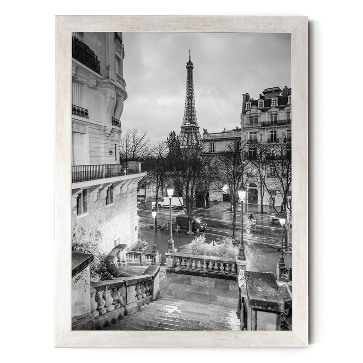 A black and white photograph of the Eiffel Tower in Paris, France, with a street scene in the foreground featuring ornate buildings and a wet, reflective surface.